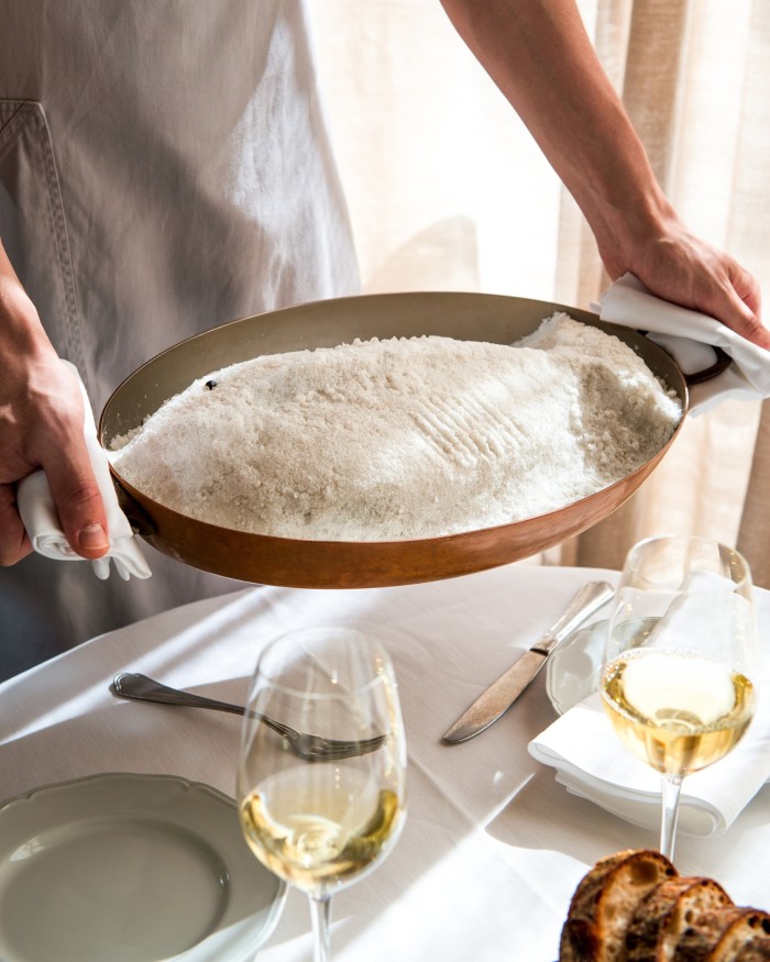 A chef's hands holding salt-roasted John Dory in a copper pan on a set table, next to two glasses of white wine